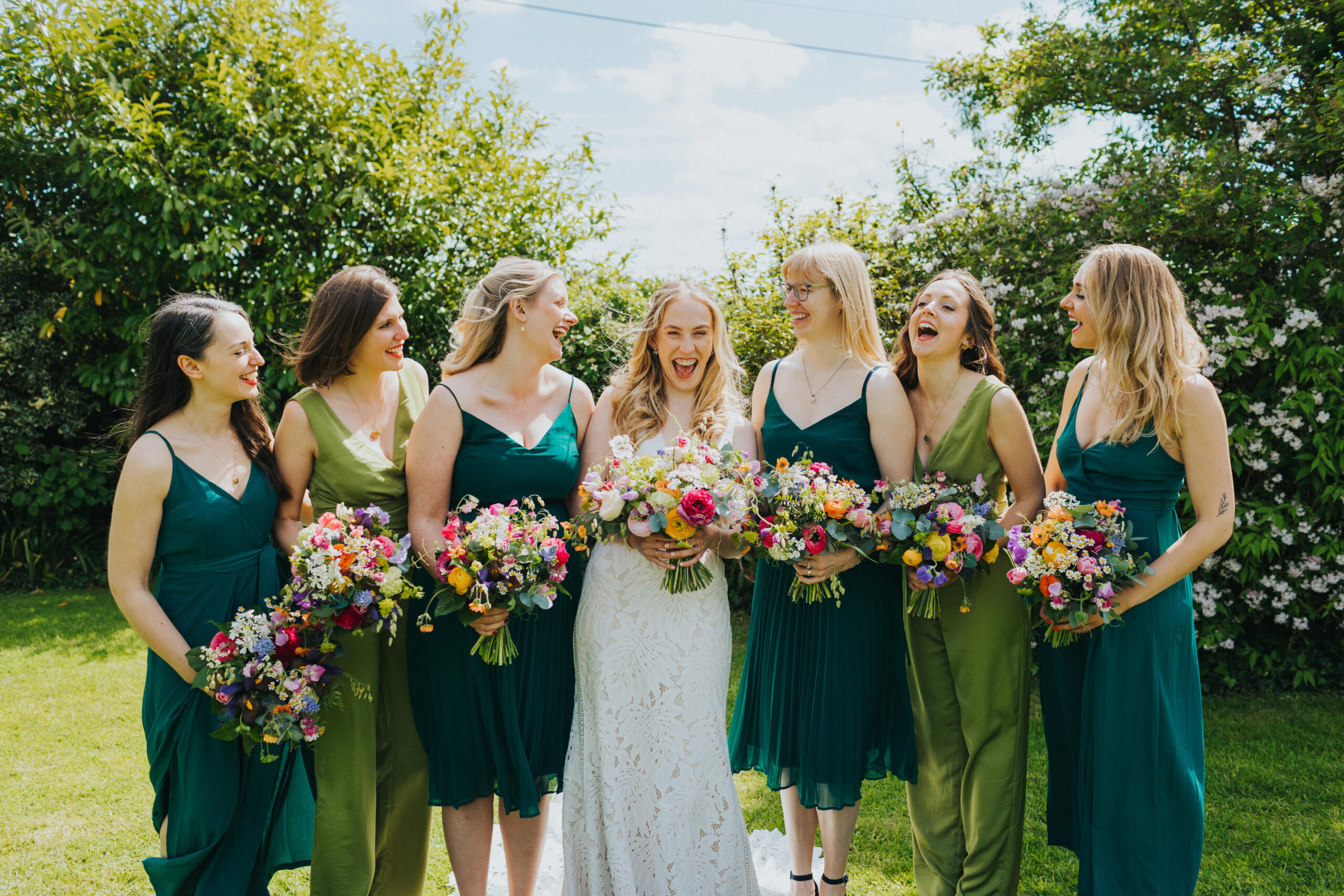 Bride with six bridesmaids, all laughing and looking at each other. The bridesmaids are all wearing different shades of green dresses.