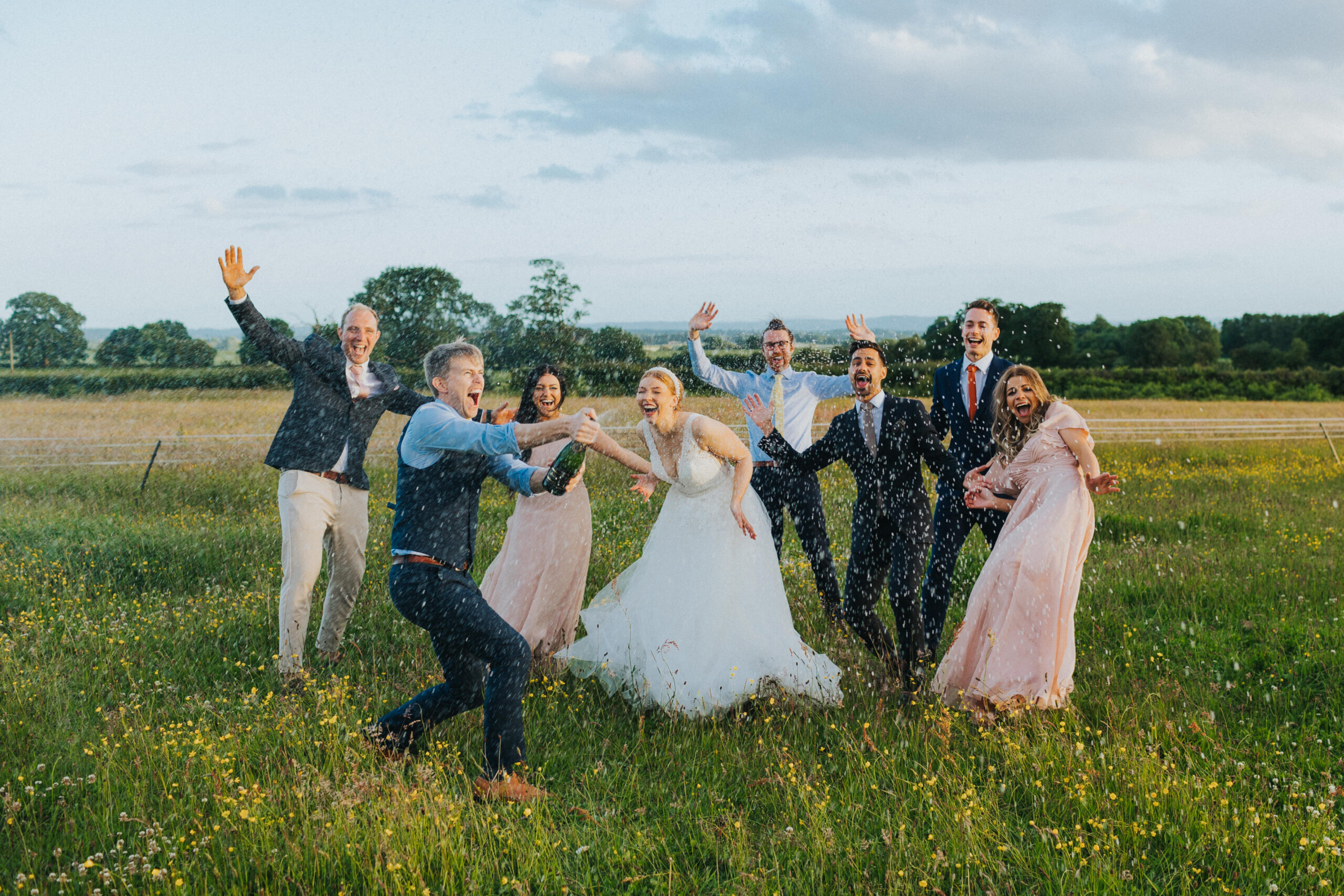 Groom, spraying a bottle of champagne, with the bride and bridal party behind him