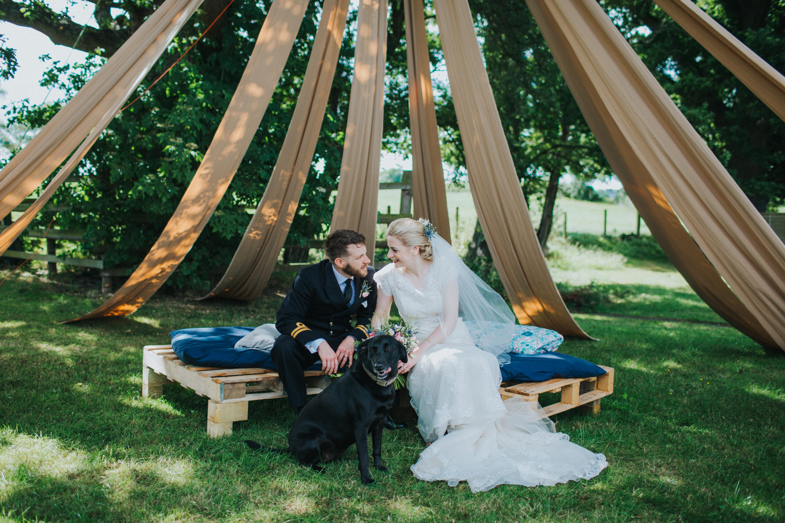bride and groom sat under tent at glamping venue with their black labrador